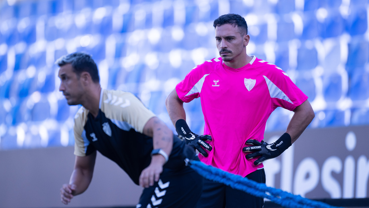 Dos futbolistas con ropa deportiva entrenan en un campo, uno con camiseta rosa y guantes y otro agachado, con gradas azules al fondo.