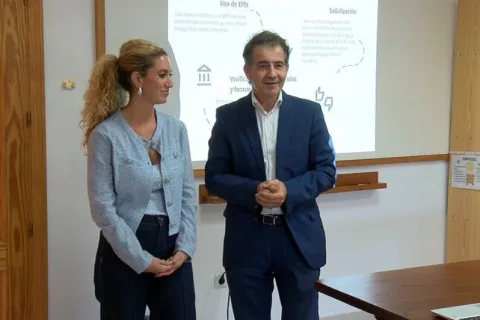 Una mujer con chaqueta azul claro y un hombre con traje azul posan de pie frente a una pantalla con texto sobre EPIs y señalización.