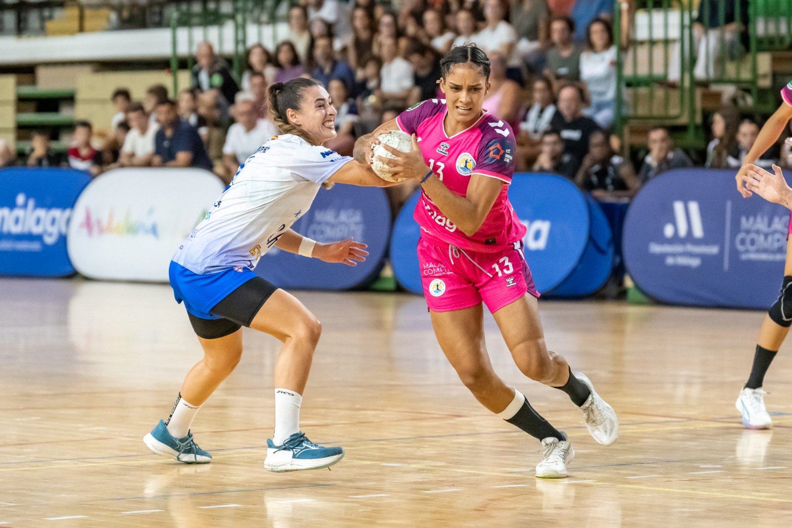 Dos jugadoras de balonmano se disputan el balón en una cancha, una con camiseta blanca y otra con equipación rosa, ante público.