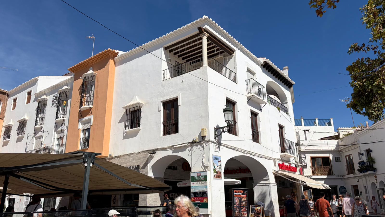 Edificio blanco de esquina con arcos y balcones, junto a otro naranja, con toldos y personas en la calle.